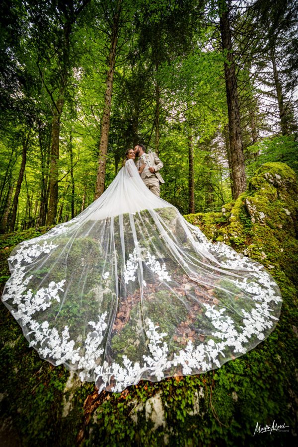 Des mariés enlacés avec un magnifique voile au premier plan, le tout en forêt